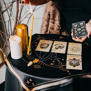May include: A black silk cloth with gold embroidery of a celestial design. Four tarot cards are laid out on the cloth, along with a lit candle, a small bottle, and two crystals. The text "100% SILK CLOTH" is printed on the cloth.