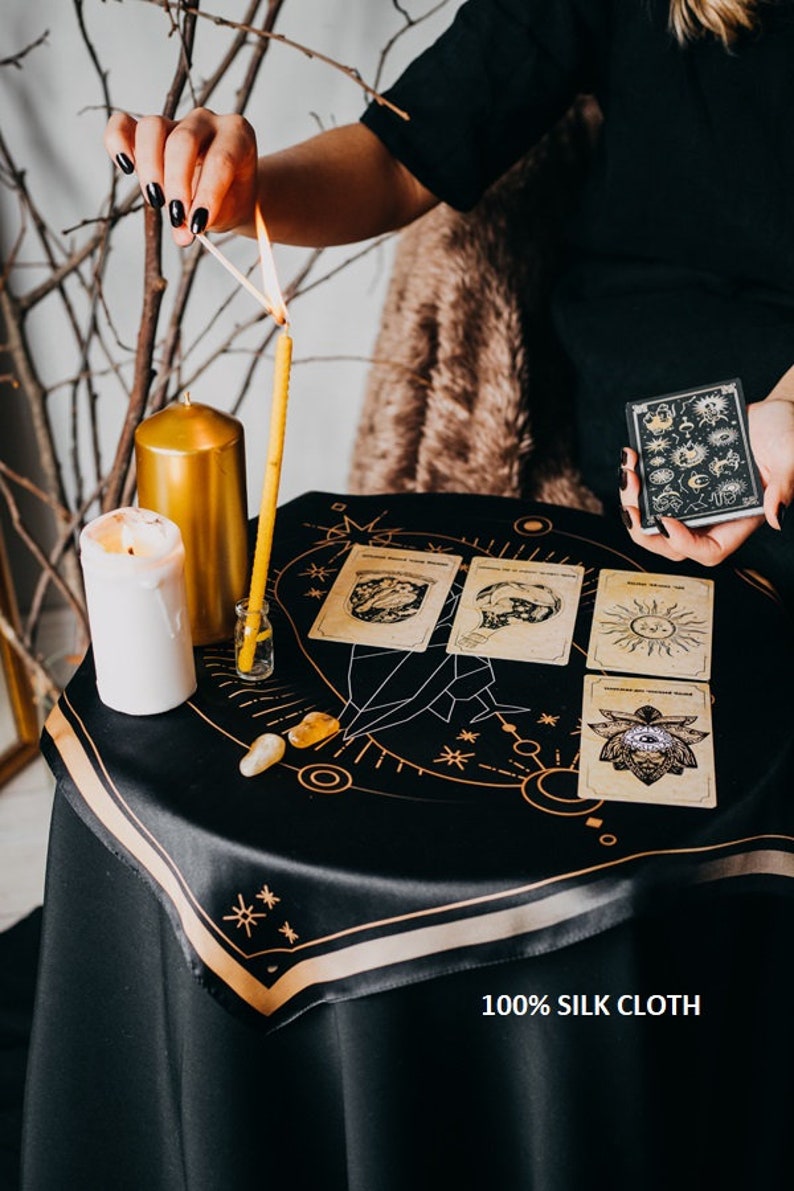 May include: A black silk cloth with gold embroidery of a celestial design. Four tarot cards are laid out on the cloth, along with a lit candle, a small bottle, and two crystals. The text "100% SILK CLOTH" is printed on the cloth.