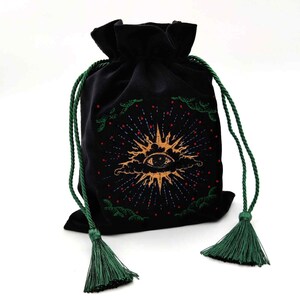 May include: Black velvet drawstring bag with a gold sun and eye design. The bag features green drawstrings and tassels. The sun design has red and blue accents. The bag is likely used for tarot cards or other spiritual items.