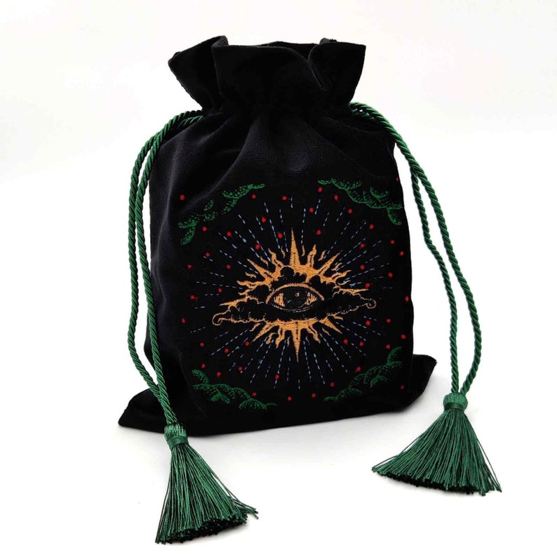 May include: Black velvet drawstring bag with a gold sun and eye design. The bag features green drawstrings and tassels. The sun design has red and blue accents. The bag is likely used for tarot cards or other spiritual items.