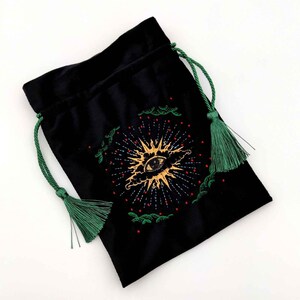 May include: Black velvet drawstring bag with a gold and red embroidered eye design. The bag has green drawstrings and tassels. The eye design is surrounded by a sunburst pattern and green embroidered accents.