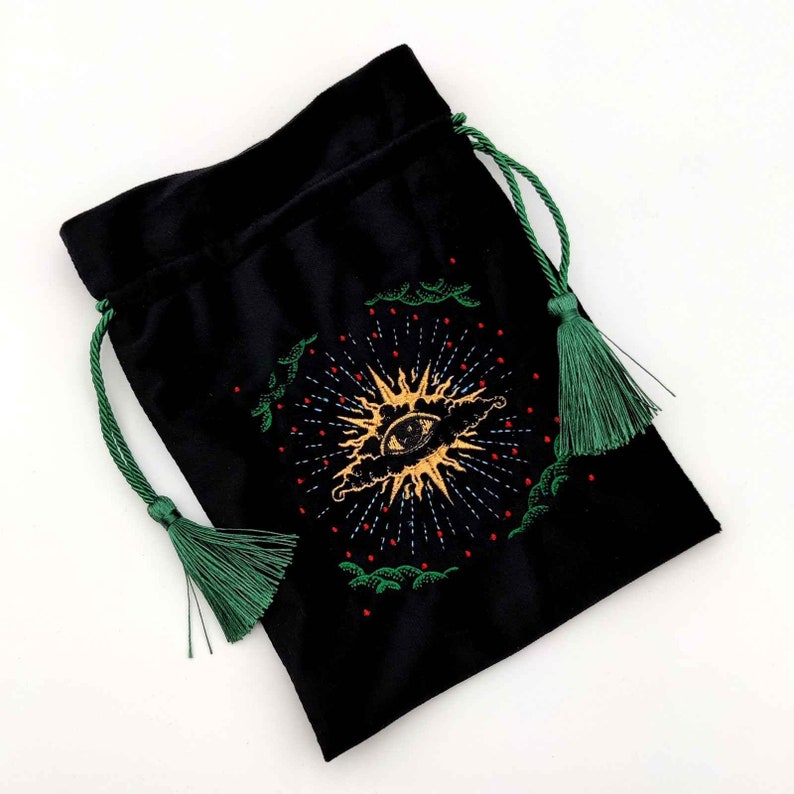 May include: Black velvet drawstring bag with a gold and red embroidered eye design. The bag has green drawstrings and tassels. The eye design is surrounded by a sunburst pattern and green embroidered accents.