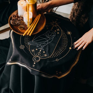 May include: A black cloth with a gold geometric owl design, surrounded by stars and moons. A wooden tray with three candles and pine cones sits on top of the cloth.
