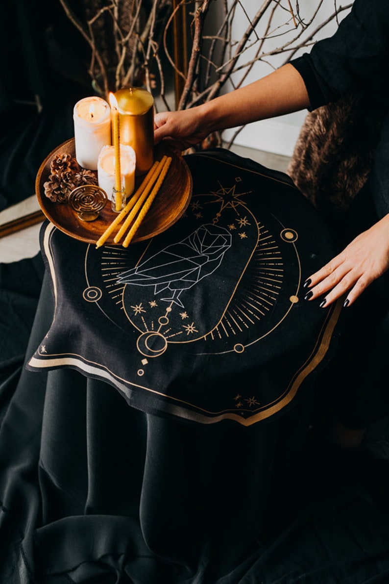 May include: A black cloth with a gold geometric owl design, surrounded by stars and moons. A wooden tray with three candles and pine cones sits on top of the cloth.