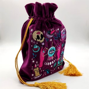 May include: A purple velvet drawstring bag with a gold tassel. The bag is embroidered with a variety of colorful images, including a skull, an eye, a potion bottle, and a candle.