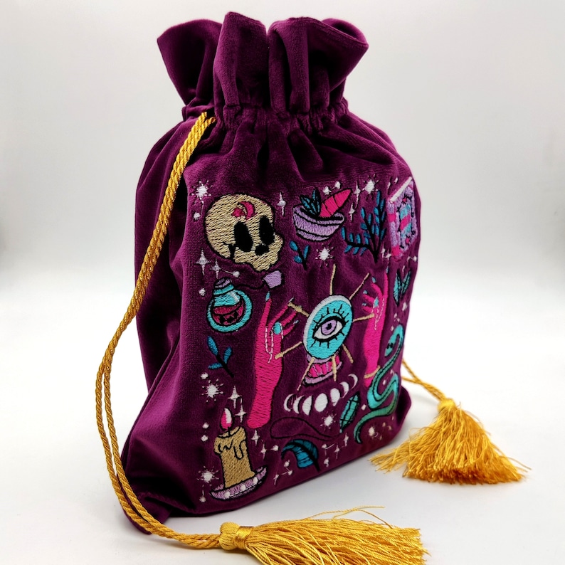 May include: A purple velvet drawstring bag with a gold tassel. The bag is embroidered with a variety of colorful images, including a skull, an eye, a potion bottle, and a candle.