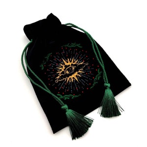 May include: Black velvet drawstring bag with an embroidered sunburst design featuring an eye. The design is in gold, red, and green. The bag has green drawstrings and tassels.