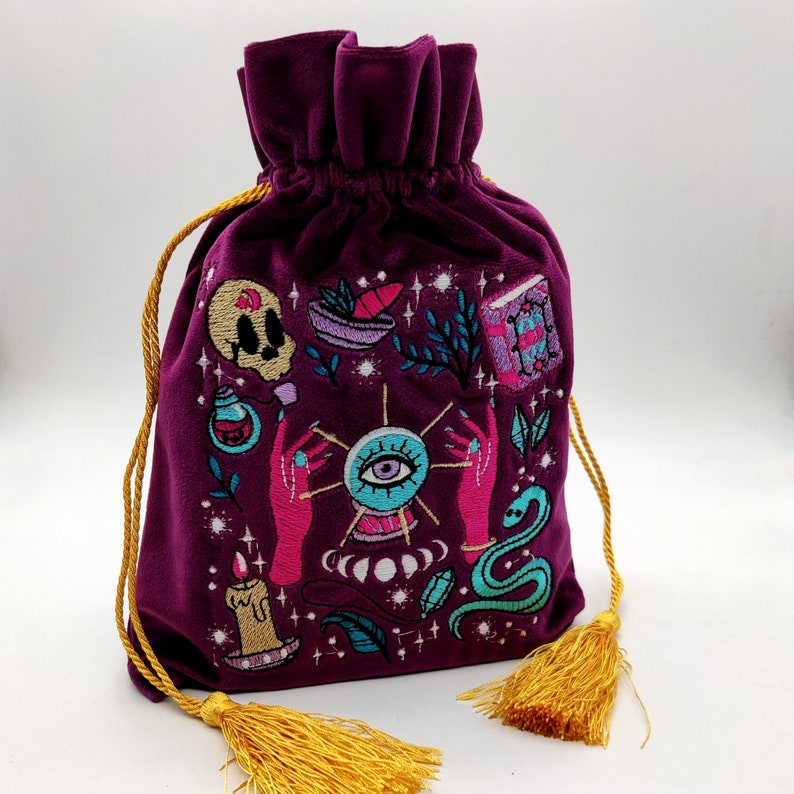 May include: A purple velvet pouch with a drawstring closure, embroidered with a mystical design featuring an eye, hands, a snake, and other symbols. The pouch has gold tassels.