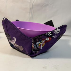 Puede incluir: Un protector de cuenco de tela morado con un estampado de Pesadilla Antes de Navidad con Jack Skellington y Sally. El protector de cuenco contiene un cuenco de plástico morado.