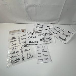 May include: A set of white labels with black handwritten text for organizing kitchen pantry items. The labels include categories like coffee, cocoa, tea, sugar, honey, cereal, pasta, snacks, nuts, granola, rice, sweets, baking powder, and spices.