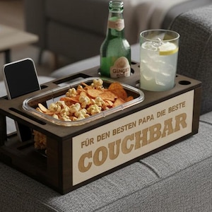 Könnte beinhalten: Ein dunkelbraunes Holz-Couch-Armtablett mit Aussparungen für ein Telefon, Snacks und Getränke. Das Tablett enthält einen transparenten Behälter mit Snacks, eine grüne Flasche und ein Glas Limonade. Der Text "COUCHBAR" ist sichtbar.