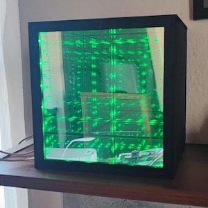 Può includere: Un cubo nero con un interno a specchio che crea un effetto di riflessione infinito. Lo specchio è pieno di luci LED verdi.
