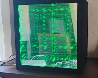 May include: A black cube with a mirrored interior that creates an infinite reflection effect. The mirror is filled with green LED lights.