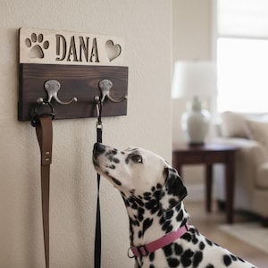 Könnte beinhalten: Ein hölzerner Hunde-Leinenhalter mit dem eingravierten Namen "DANA", zusammen mit einem Pfotenabdruck und einem Herz. Der Halter hat zwei Metallhaken und ist an einer Wand montiert. Eine braune Lederleine und eine schwarze Leine hängen an den Haken. Ein Dalmatiner mit rosa Halsband blickt zu den Leinen auf.