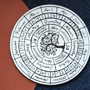 Könnte beinhalten: Ein weißer, runder Holzkalender mit schwarzen Markierungen. Der Kalender zeigt die Monate des Jahres, die Wochentage und ein Baumdesign in der Mitte. Der Text auf dem Kalender beinhaltet "Montag", "Dienstag", "Mittwoch", "Donnerstag", "Freitag", "Samstag", "Sonntag", "Ja", "Nein", "Vielleicht", "nochmal versuchen", und "Januar", "Februar", "März", "April", "Mai", "Juni", "Juli", "August", "September", "Oktober", "November", "Dezember".