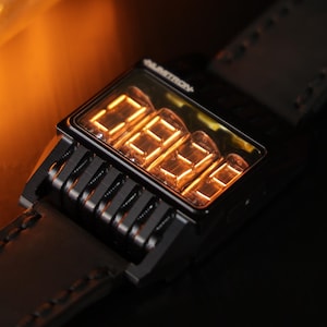May include: A black digital wristwatch with a leather strap. The watch face displays illuminated orange numbers. A glowing light bulb is in the background. The watch has a unique, retro-futuristic design.