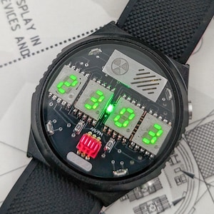 Könnte beinhalten: Schwarze Armbanduhr mit einem Zifferblatt mit Leiterplatte, das grüne digitale Zahlen anzeigt. Die Uhr hat ein schwarzes Armband und eine rote Komponente. Das Zifferblatt enthält den Text "AM/PM".