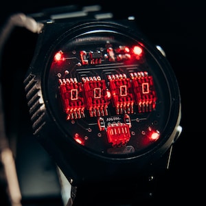 May include: Black wristwatch with a unique circuit board design. The watch face features glowing red lights and a digital display. The watchband is black and metallic. The overall aesthetic is futuristic and tech-inspired.