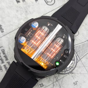 Könnte beinhalten: Eine schwarze Armbanduhr mit einem klaren Zifferblatt und einem einzigartigen Design. Das Zifferblatt der Uhr verfügt über eine digitale Anzeige mit orange leuchtenden Zahlen und einer silbernen Metallstange. Die Uhr hat ein schwarzes Kautschukband.