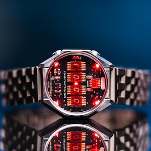 May include: A futuristic wristwatch with a silver-tone case and a black face displaying red digital readouts. The watchband is a matching silver-tone, with a geometric pattern. The watch is reflected on a blue surface.