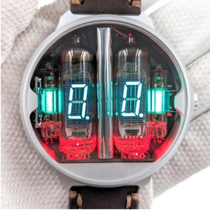 May include: A silver Nixie tube watch with a brown leather strap. The watch face displays digital numbers in green and red, with the brand name "NIXDID" visible. The back of the watch has a circuit board design and the word "SPACE".