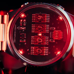 May include: Close-up of a futuristic wristwatch with a silver band and a dark red face. The watch face displays illuminated red digital numbers and circuit board details. The watch is set against a dark background.