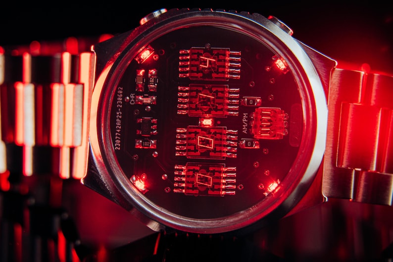 May include: Close-up of a futuristic wristwatch with a silver band and a dark red face. The watch face displays illuminated red digital numbers and circuit board details. The watch is set against a dark background.
