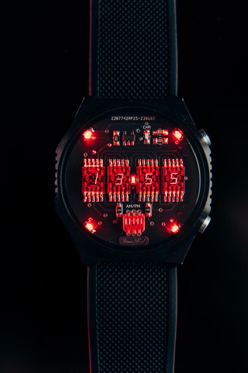 May include: Black digital watch with a circuit board design. The watch face displays the time in red LED numerals. The watch band is black and textured. The watch has the text "Time Pod" on the face.