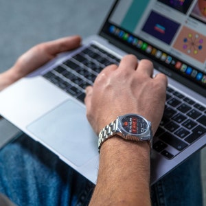 May include: A silver wristwatch with a black face and red digital display is worn on a wrist. The watch has a stainless steel band. A laptop computer is open in the background, displaying a colorful website.