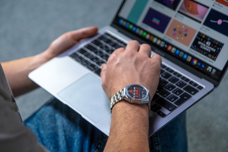 May include: A silver wristwatch with a black face and red digital display is worn on a wrist. The watch has a stainless steel band. A laptop computer is open in the background, displaying a colorful website.