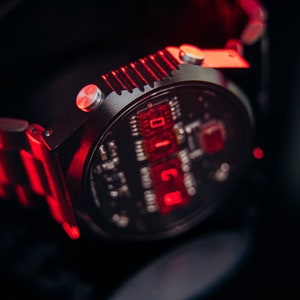 May include: Close-up of a black wristwatch with a digital display showing red numerals. The watch has a metallic band and a circular face with intricate internal components visible. The watch is illuminated with red light.