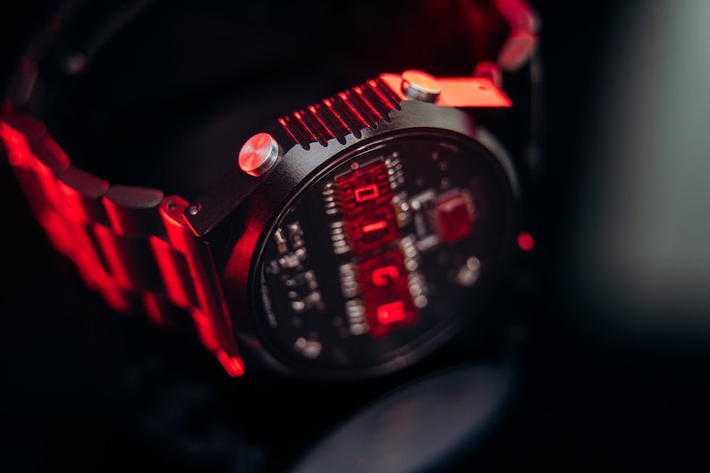 May include: Close-up of a black wristwatch with a digital display showing red numerals. The watch has a metallic band and a circular face with intricate internal components visible. The watch is illuminated with red light.
