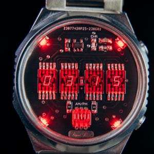 May include: A close-up of a digital wristwatch with a silver band. The watch face displays the time in bright red LED lights against a black background. The watch has a unique circuit board design.