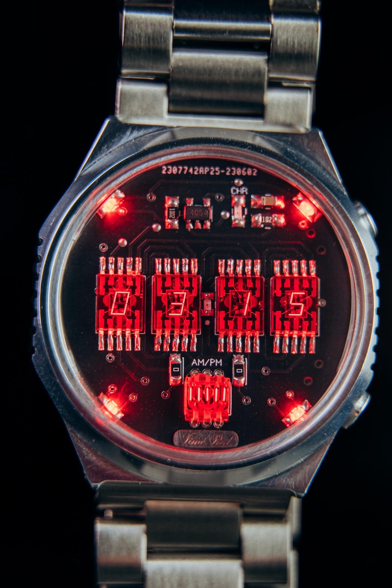 May include: A close-up of a digital wristwatch with a silver band. The watch face displays the time in bright red LED lights against a black background. The watch has a unique circuit board design.