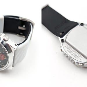 May include: A silver wristwatch with a white and black band. The watch face has a digital display with red numbers. The back of the watch has a silver case with black text.