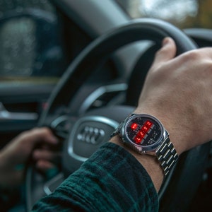 May include: A silver wristwatch with a digital red display showing the time. The watch is on a person's wrist, with a silver band. The person is holding a steering wheel, wearing a green and black plaid shirt.