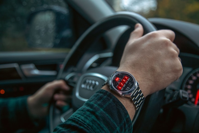 May include: A silver wristwatch with a digital red display showing the time. The watch is on a person's wrist, with a silver band. The person is holding a steering wheel, wearing a green and black plaid shirt.