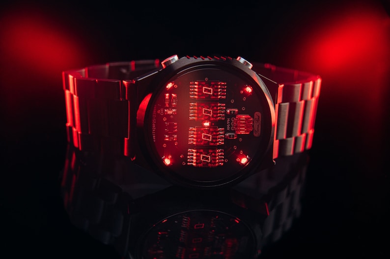 May include: A futuristic wristwatch with a black face and a metallic band. The watch face displays red illuminated digital components. The watch is set against a dark background with red lighting, creating a high-tech aesthetic.