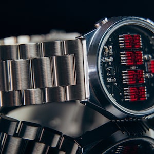 May include: A silver-toned wristwatch with a stainless steel band. The watch face reveals a circuit board design with red digital displays. The watch is displayed on a reflective surface, creating a mirrored effect.