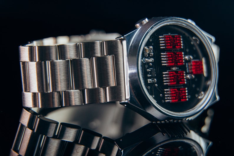 May include: A silver-toned wristwatch with a stainless steel band. The watch face reveals a circuit board design with red digital displays. The watch is displayed on a reflective surface, creating a mirrored effect.