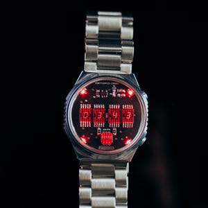 May include: A silver-toned wristwatch with a digital display. The watch face features red LED numerals and a circuit board design. The watch has a metal band with a clasp closure, and the time displayed is 03:43.