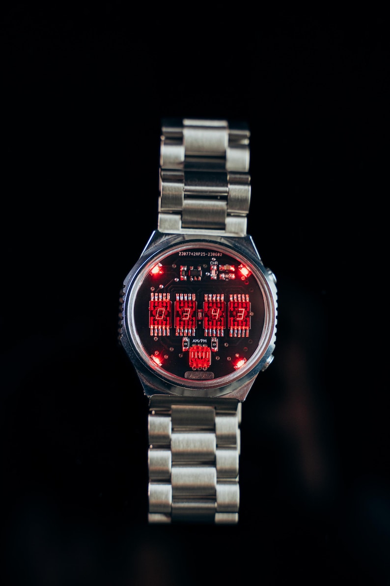 May include: A silver-toned wristwatch with a digital display. The watch face features red LED numerals and a circuit board design. The watch has a metal band with a clasp closure, and the time displayed is 03:43.
