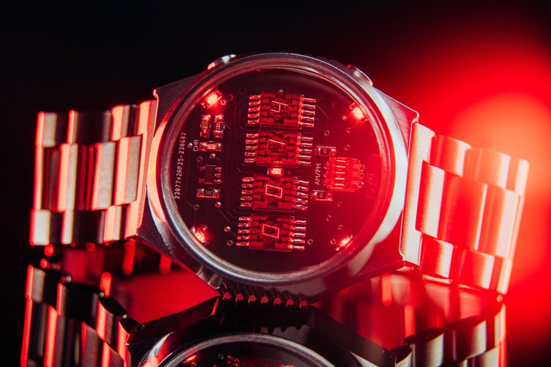 May include: A silver wristwatch with a black face and red digital display. The watch has a metal band and is illuminated with red light. The watch face displays the time in a digital format. The watch is a retro-futuristic design.
