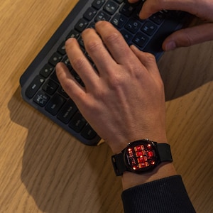 May include: A black smartwatch with a red digital display is worn on a wrist. The watch is on a person's wrist as they type on a black keyboard. The keyboard is on a light brown wooden surface.
