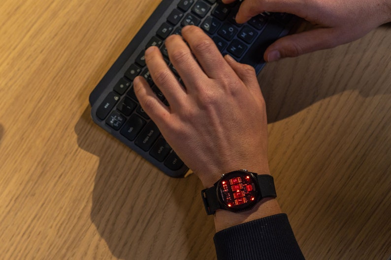 May include: A black smartwatch with a red digital display is worn on a wrist. The watch is on a person's wrist as they type on a black keyboard. The keyboard is on a light brown wooden surface.