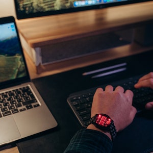 May include: A black digital watch with a red LED display is worn on a wrist. The watch is on a black desk next to a laptop and a keyboard. The watch has a metal band. The person is wearing a plaid shirt.