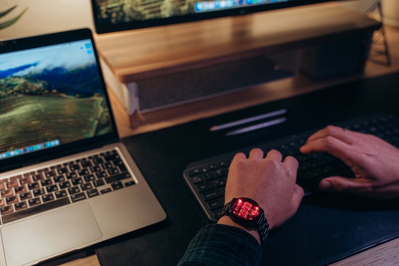 May include: A black digital watch with a red LED display is worn on a wrist. The watch is on a black desk next to a laptop and a keyboard. The watch has a metal band. The person is wearing a plaid shirt.