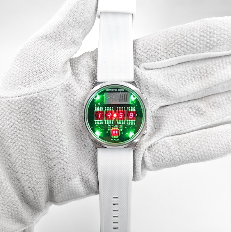 May include: A silver wristwatch with a white band and a green and red digital display. The display shows the time as 1:45:58. The watch has the text "24/7 TEMPO - 32847" and "MEXICO" printed on the face.