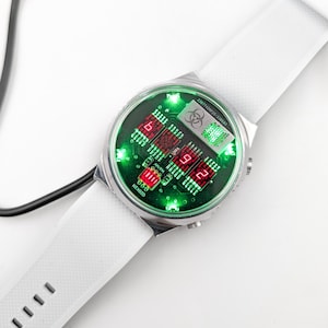 May include: A white wristwatch with a silver face and a green and red digital display. The watch face has a biohazard symbol and the text "24/7 Virus-Current".