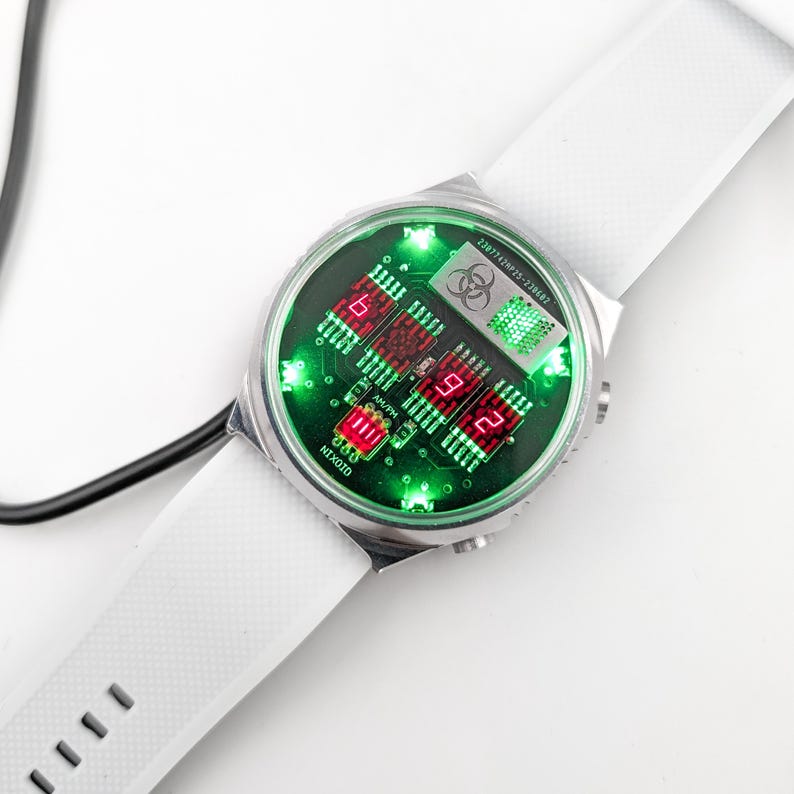 May include: A white wristwatch with a silver face and a green and red digital display. The watch face has a biohazard symbol and the text "24/7 Virus-Current".